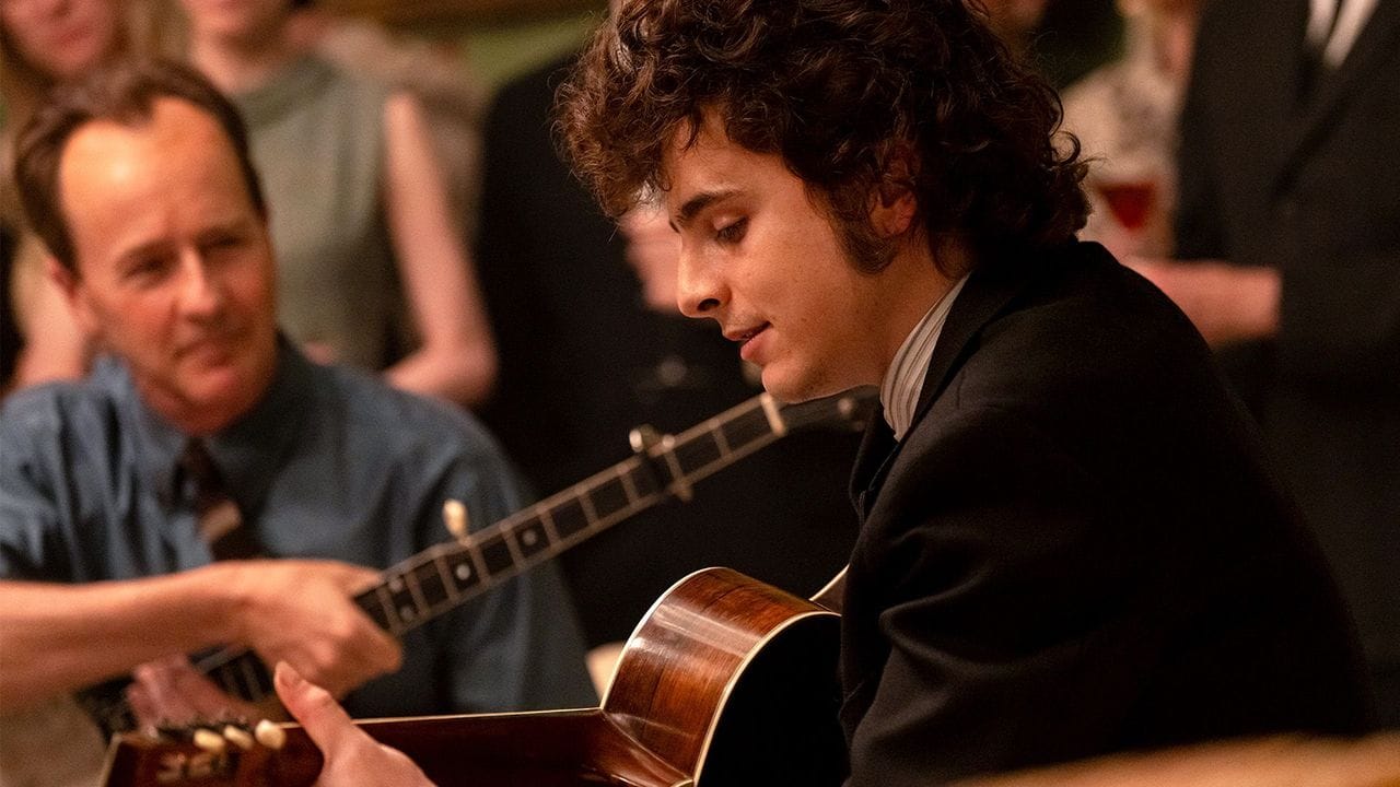 A still of Timothee Chalamet as Bob Dylan, playing guitar, with Edward Norton in the background looking insane with the most tragic receding hairline I've seen on film in a while