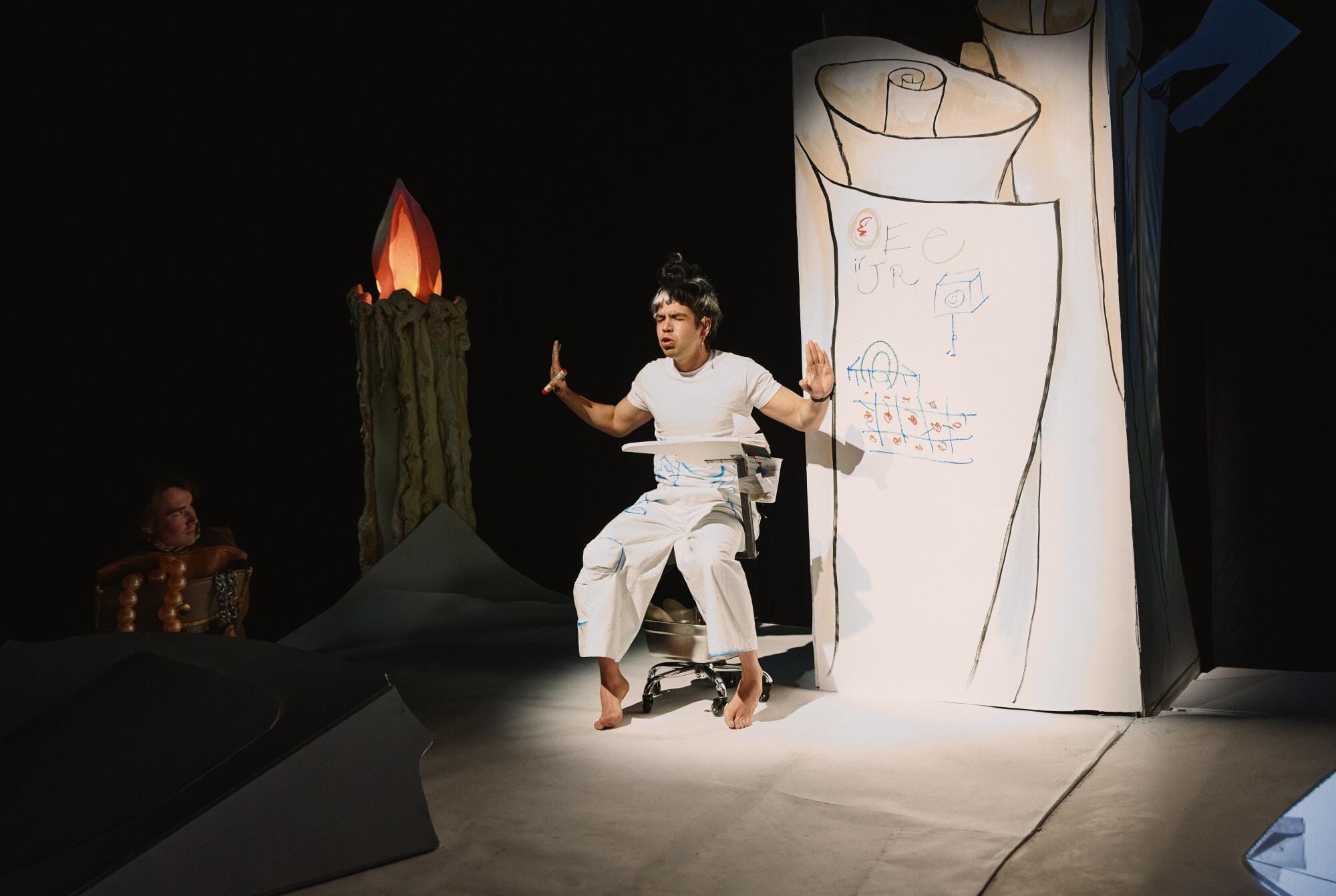 A photo of Julio Torres onstage during a performance of Color Theores — he's in an all-white outfit, in front of an all-white gigantic sheet of paper, on which we can see different colors and letters he's scribbled in colored markers
