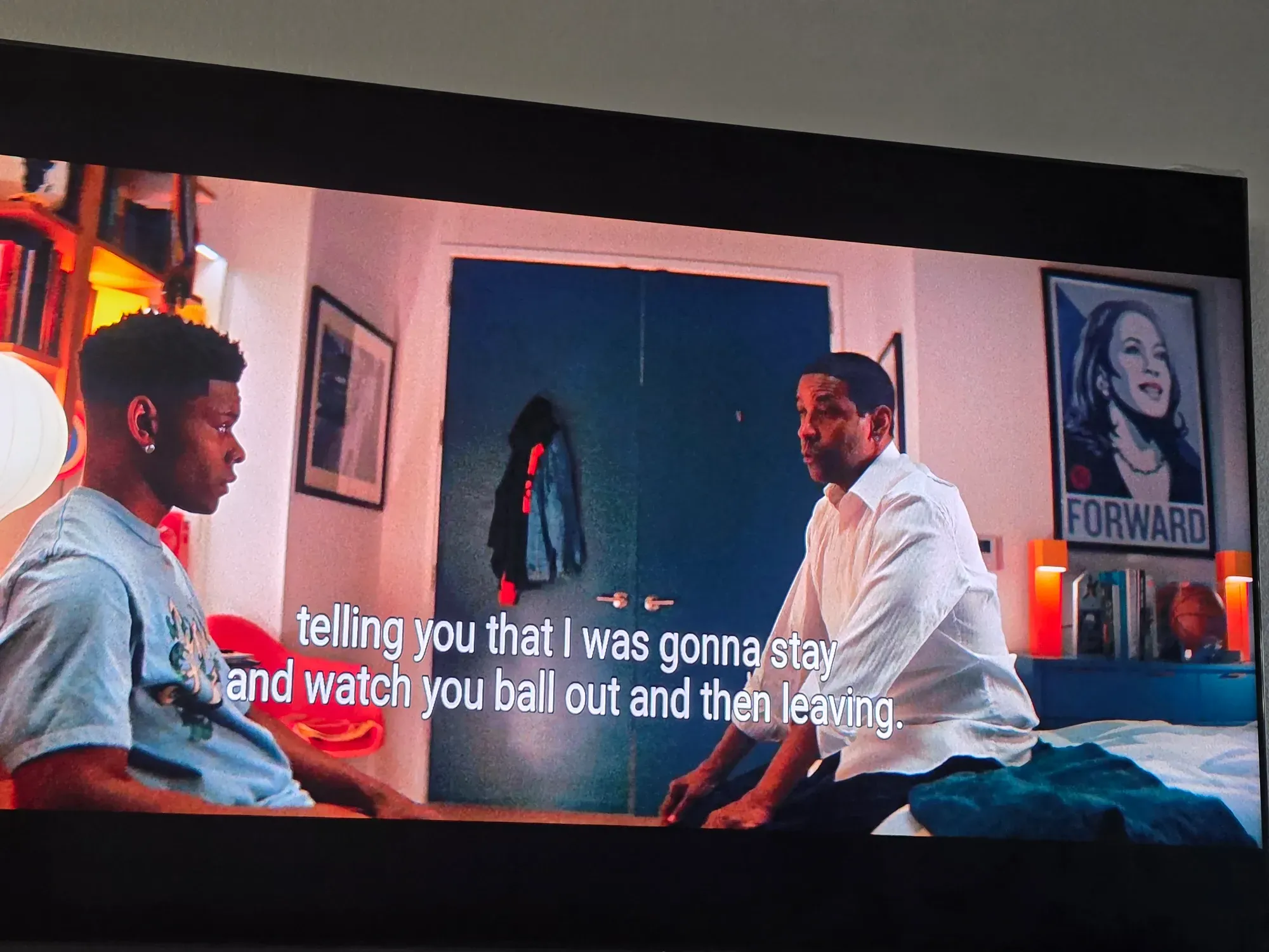 A still from the movie, showing Denzel Washington in conversation with his son, while behind them and hanging over the entire scene is a completely insane Kamala Harris campaign poster, framed and hung on the wall like it's a movie poster or a sports star