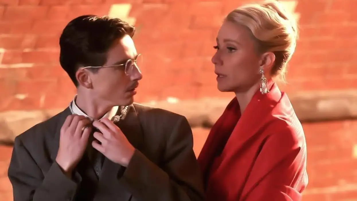 A still from Marty Supreme, showing Gwyneth Paltrow looking gorgeous in a red dress and dangly earrings, talking to Timothee Chalamet who is in a hilariously oversized suit that he's positively swimming in